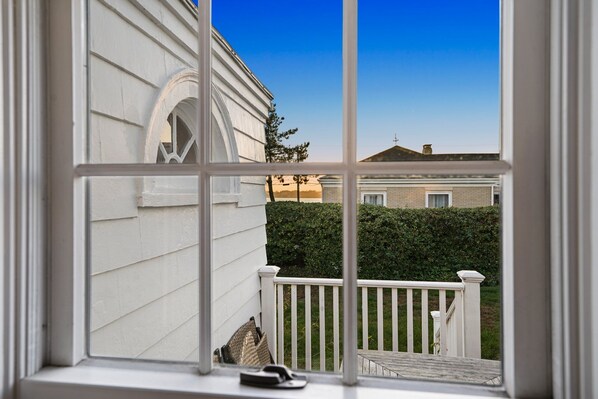 A view from a window with a white railing, a white building with a brick section, and a hedge in the background.