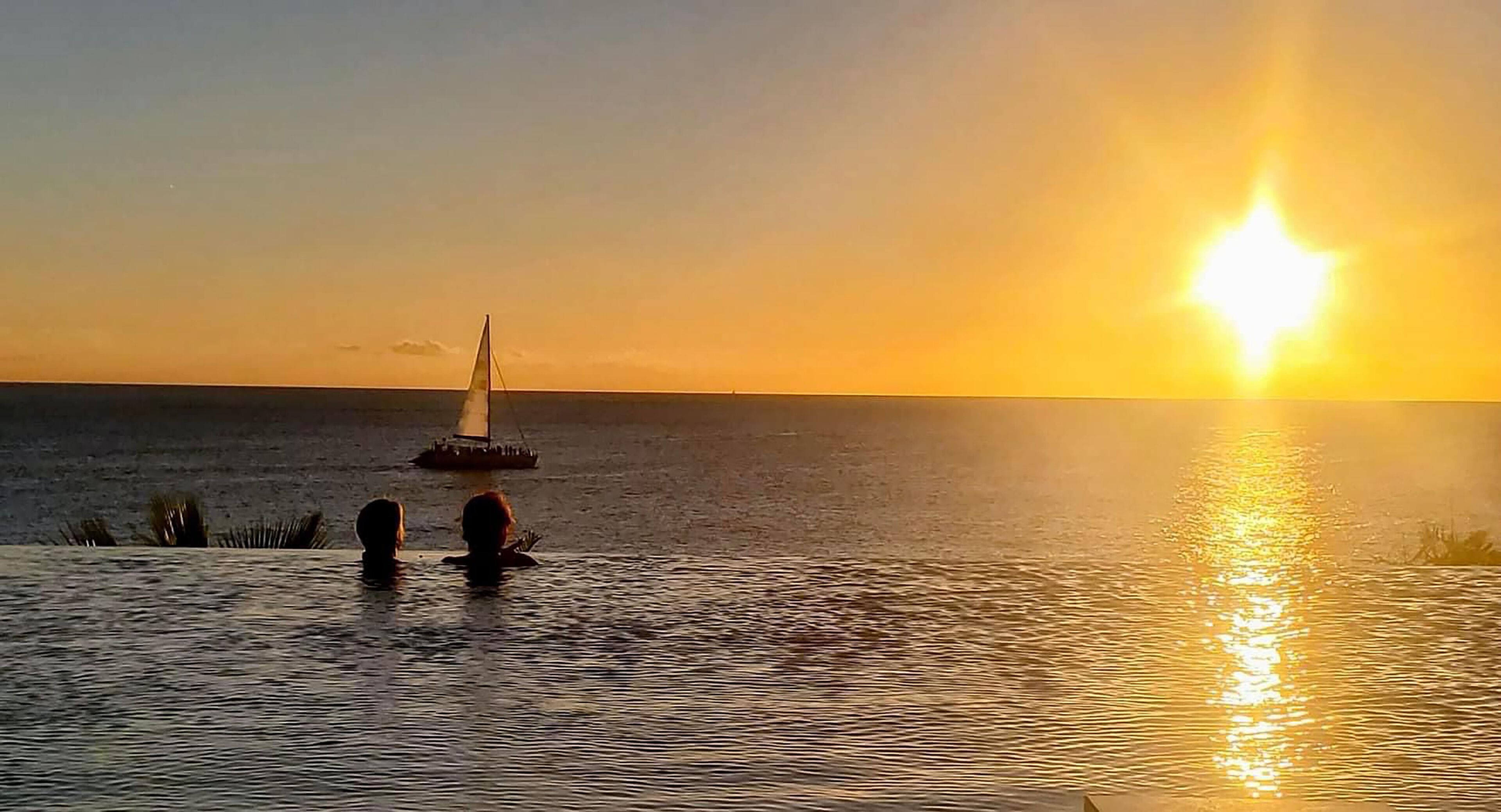 Un coucher de soleil sur une étendue d’eau calme, avec un voilier et deux personnes au premier plan.