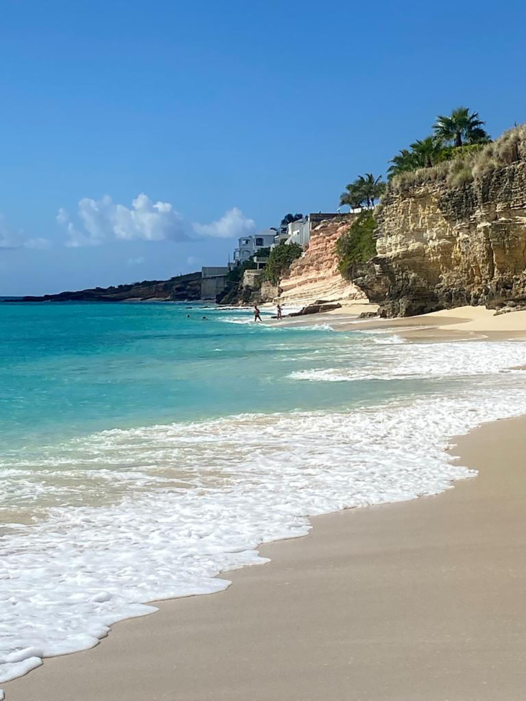 A beach with clear turquoise water, a sandy shore, and a rocky cliff with buildings on top.