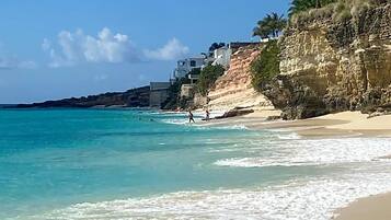 A beach with clear turquoise water, a sandy shore, and a rocky cliff with buildings on top.