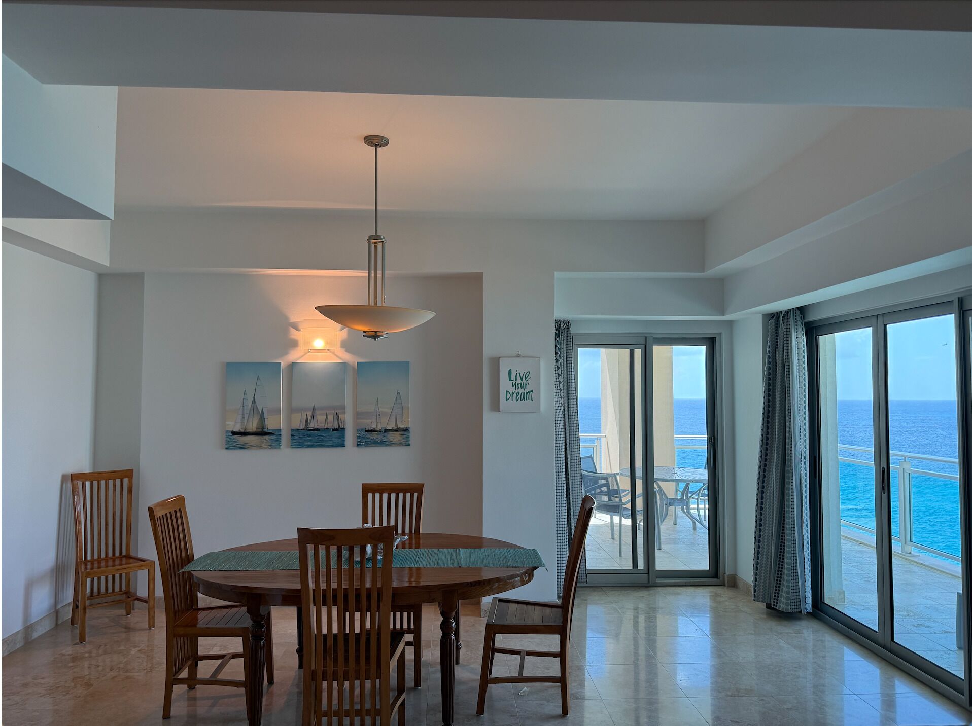 A dining area with a round table and chairs, a pendant light, and a view of the ocean through a sliding glass door.