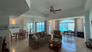 A spacious living area with a large window overlooking the sea, a ceiling fan, and a dining area with wooden furniture. - The Cliffs 2BR 2.5BA Apt Private Beach Sleeps 5
Luxury Oceanfront Apartment (Cupecoy)