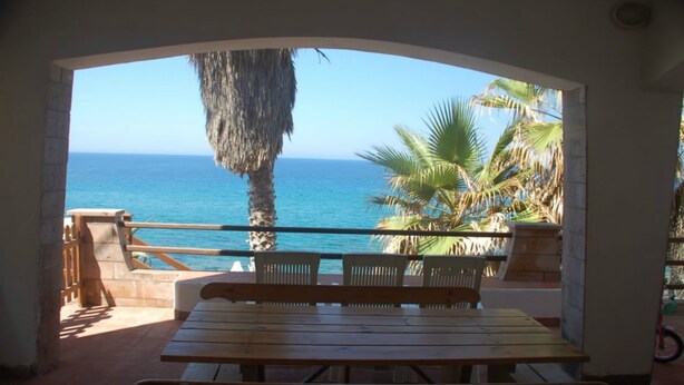 A wooden bench and table in a covered outdoor area with a view of the sea and palm trees. - Villa Bosa, Waterfront apartment in Bosa area, 10 meters from beach WI (trasnuraghes)