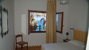 A bedroom with a bed, a chair, a nightstand, a lamp, a window with a view of the ocean and palm trees, and a curtain. - Villa Bosa, Waterfront apartment in Bosa area, 10 meters from beach WI (trasnuraghes)