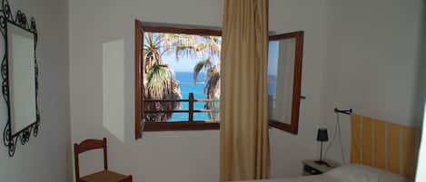 A bedroom with a bed, a chair, a nightstand, a lamp, a window with a view of the ocean and palm trees, and a curtain.