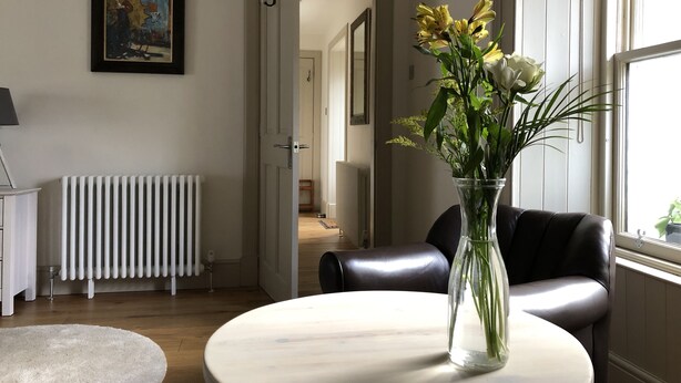 A room with a round table, a vase with flowers, a radiator, a framed picture on the wall, and a window with blinds.