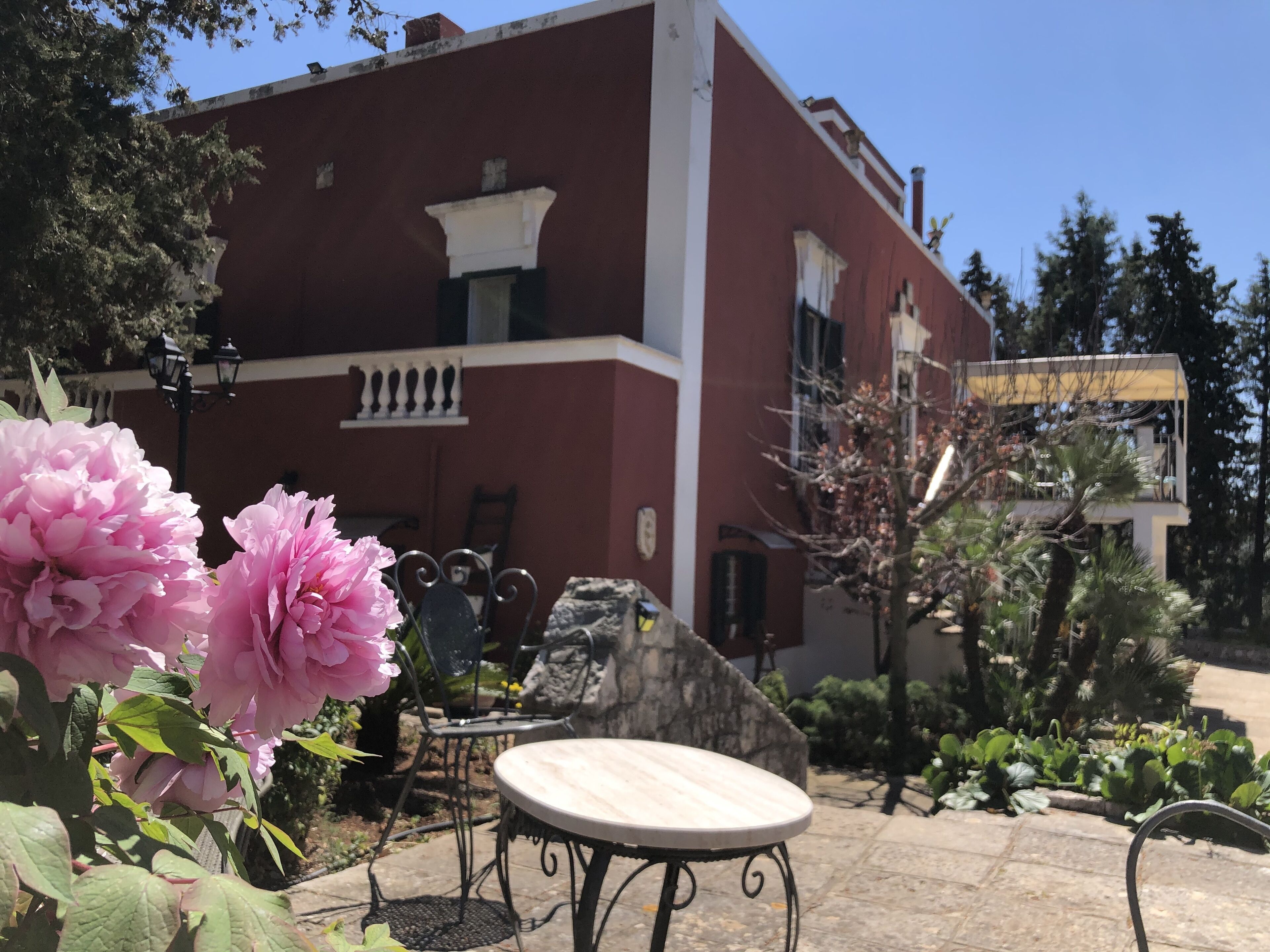 A red building with a balcony, a round table, and chairs in a garden area.