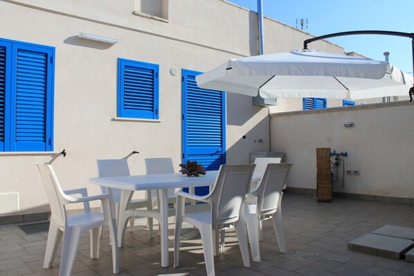 An outdoor patio with a table and chairs, a white umbrella, and blue shutters on the building.