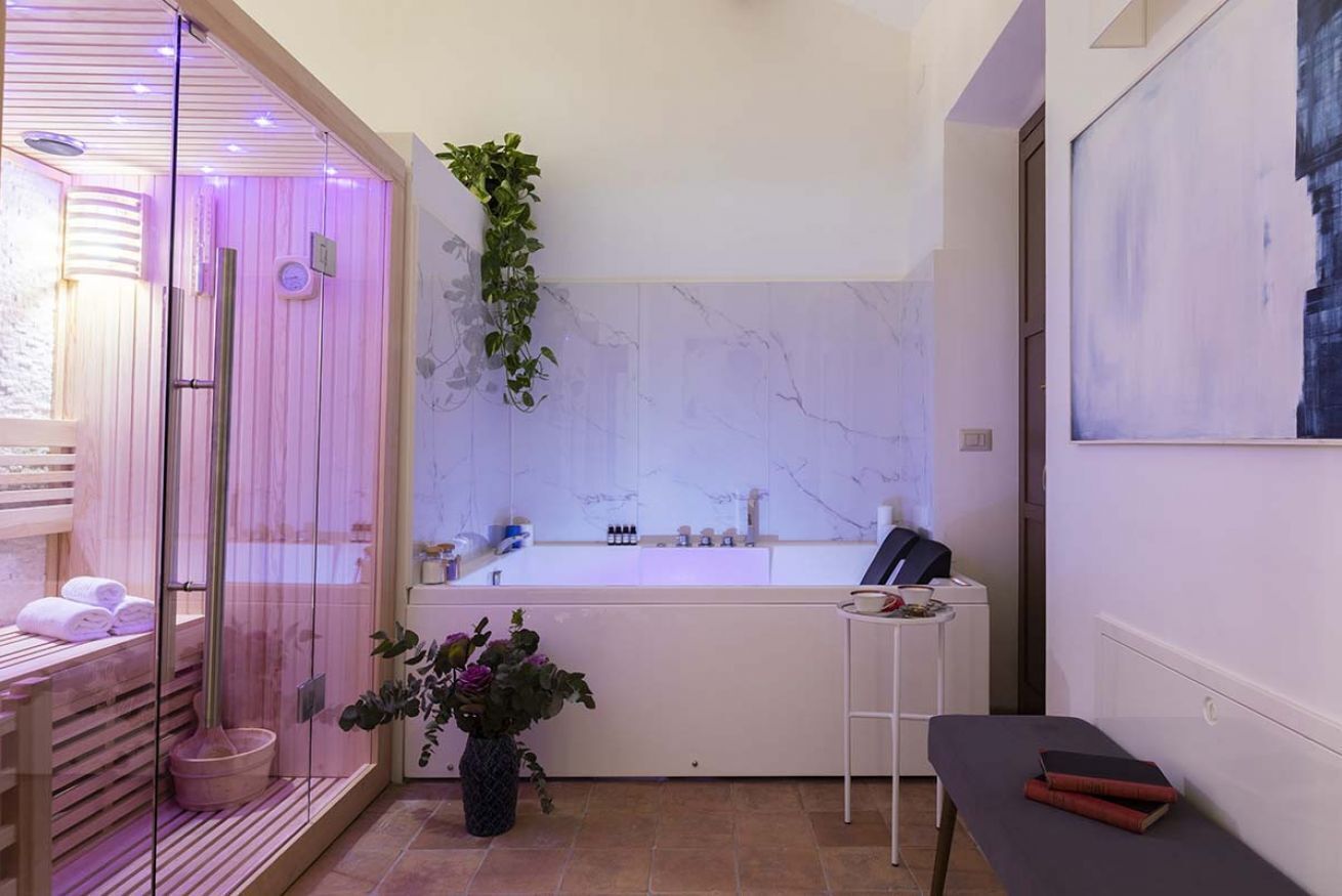 Ein moderner Spa-Bereich mit einer gläsernen Sauna, einer Wand mit Marmorplatten, einer Holzbank mit Handtüchern, einem kleinen Tisch mit Büchern und einer Topfpflanze.