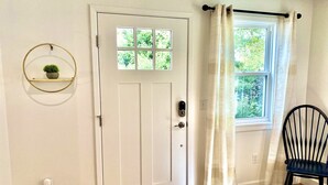 A room with a white door, a window with curtains, a wooden floor, a black chair, and a wall-mounted shelf with a plant.