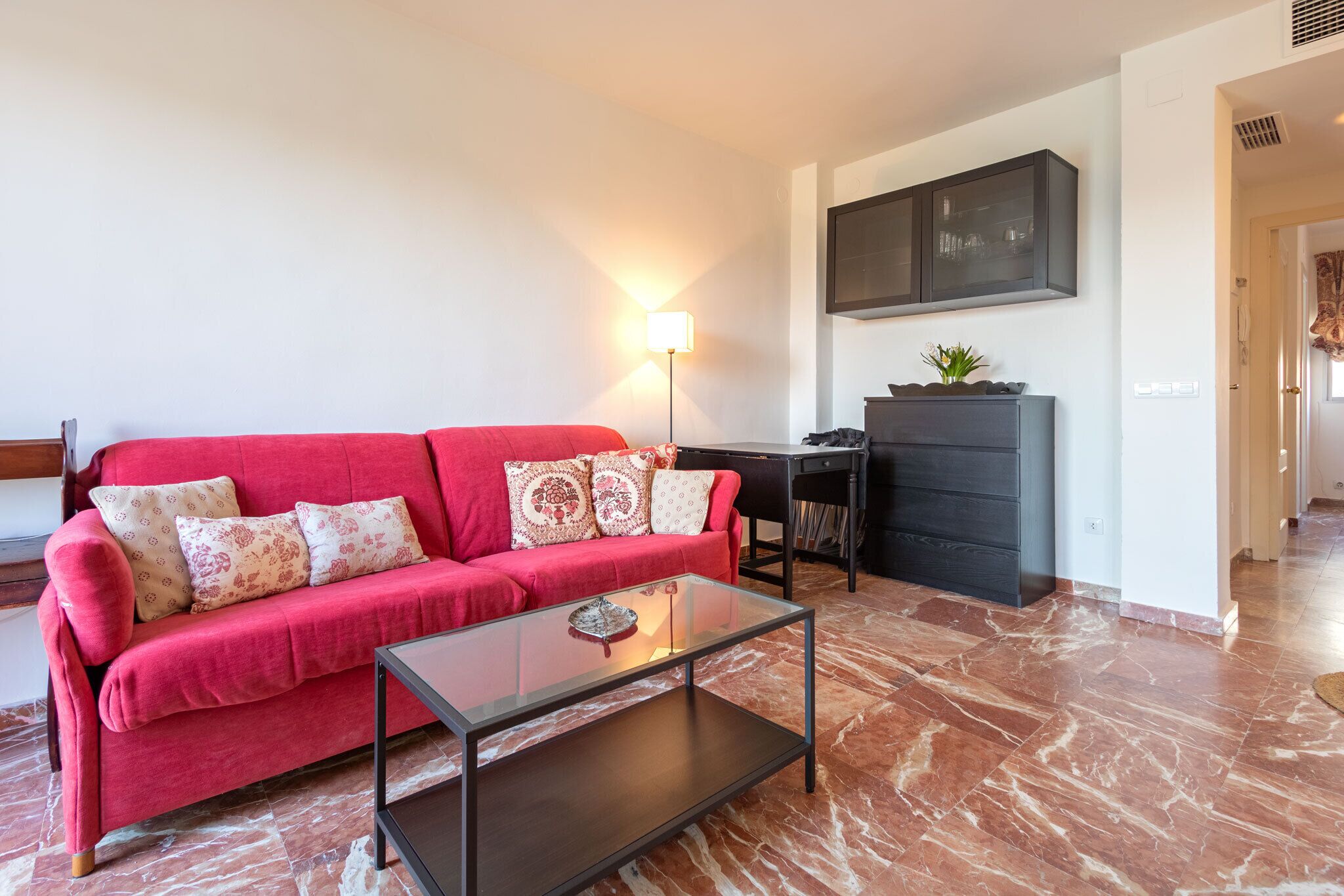 A living room with a red sofa, a glass coffee table, a desk, and a cabinet.