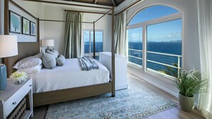 Un letto a baldacchino con vista sull'oceano, un'ampia finestra con tende e una ventola a soffitto.