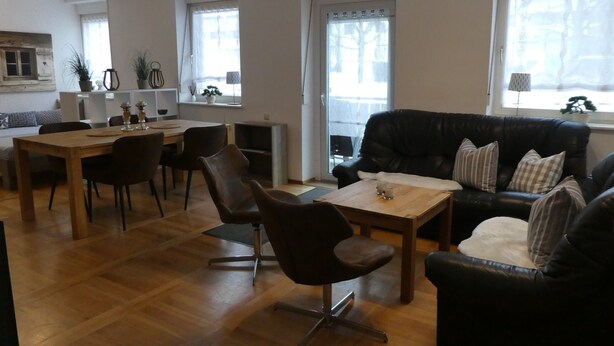 Un vaste espace de vie comprenant une table à manger en bois, un canapé en cuir noir et un fauteuil en cuir noir. On y trouve des fenêtres avec des rideaux, une cheminée et, au fond, un coin cuisine.