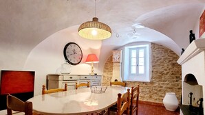 A dining area with a round table, wooden chairs, a hanging light fixture, a clock, and a stone wall. - Quality charming house classified 3* with private pool and air conditionning (La Caunette)