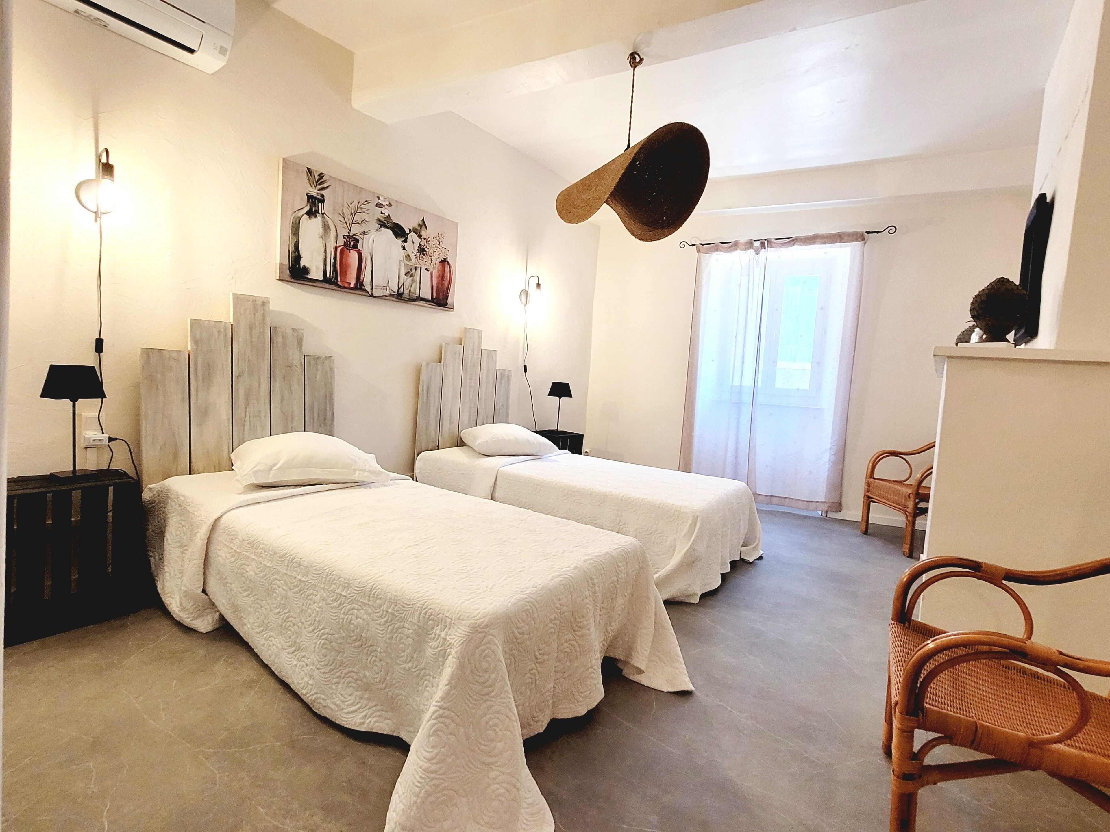 Une chambre d’hôtel avec deux lits, un chapeau accroché, un tableau et une chaise en osier.