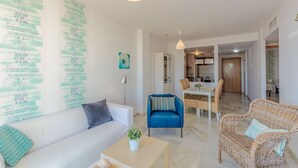 Ein modernes Wohnzimmer mit einem weißen Sofa, einem blauen Sessel und einem Korbstuhl. Es gibt einen Essbereich mit Tisch und Stühlen, und im Hintergrund eine Küche.
