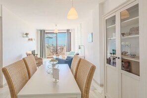 Un comedor con una mesa blanca y sillas de mimbre, un aparador de vidrio con un cartel de 'Welcome' y vistas a través de la puerta de cristal.