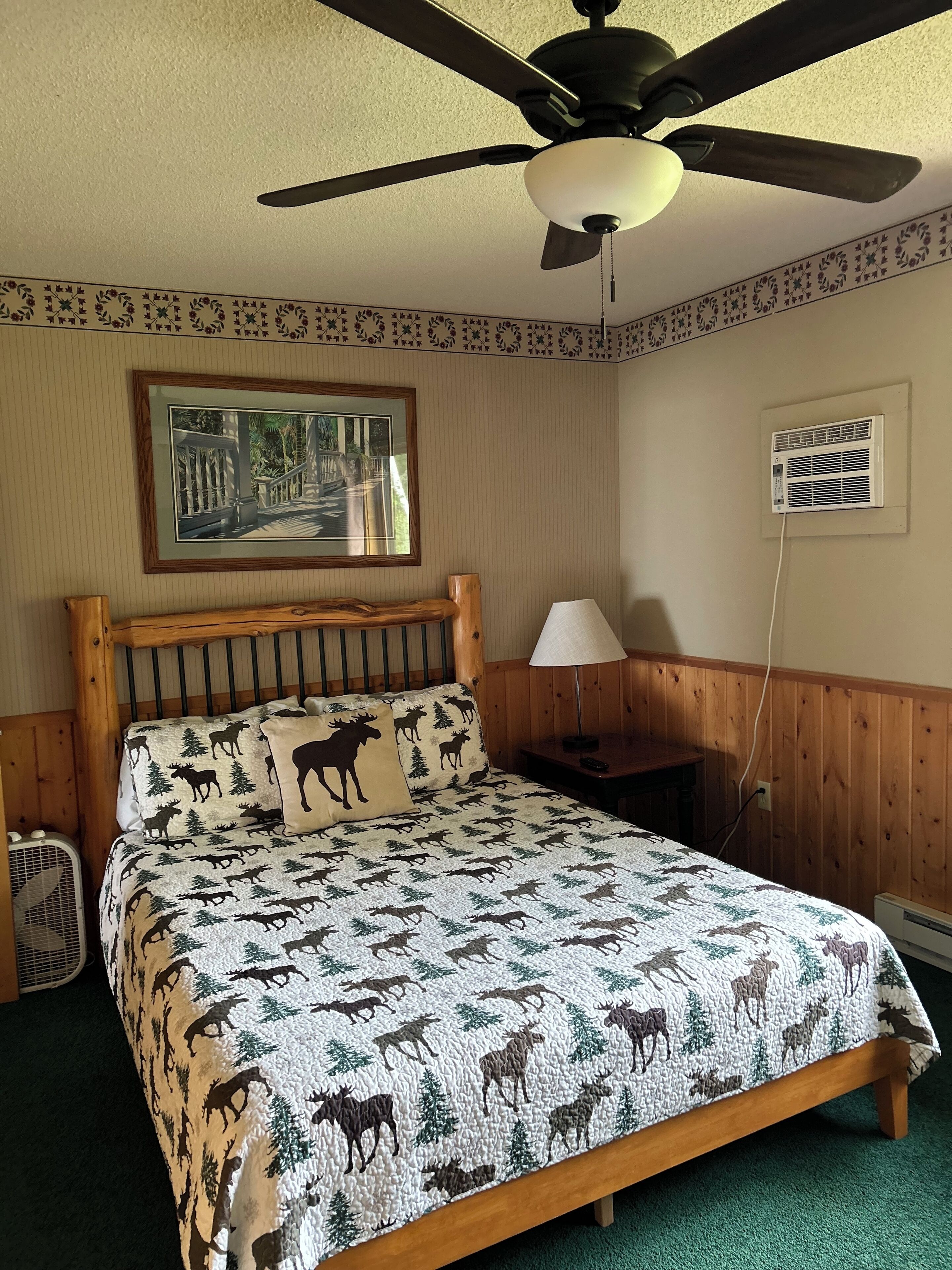 A bedroom with a bed featuring a moose print bedspread, a ceiling fan, a framed picture on the wall, and a bedside table with a lamp.