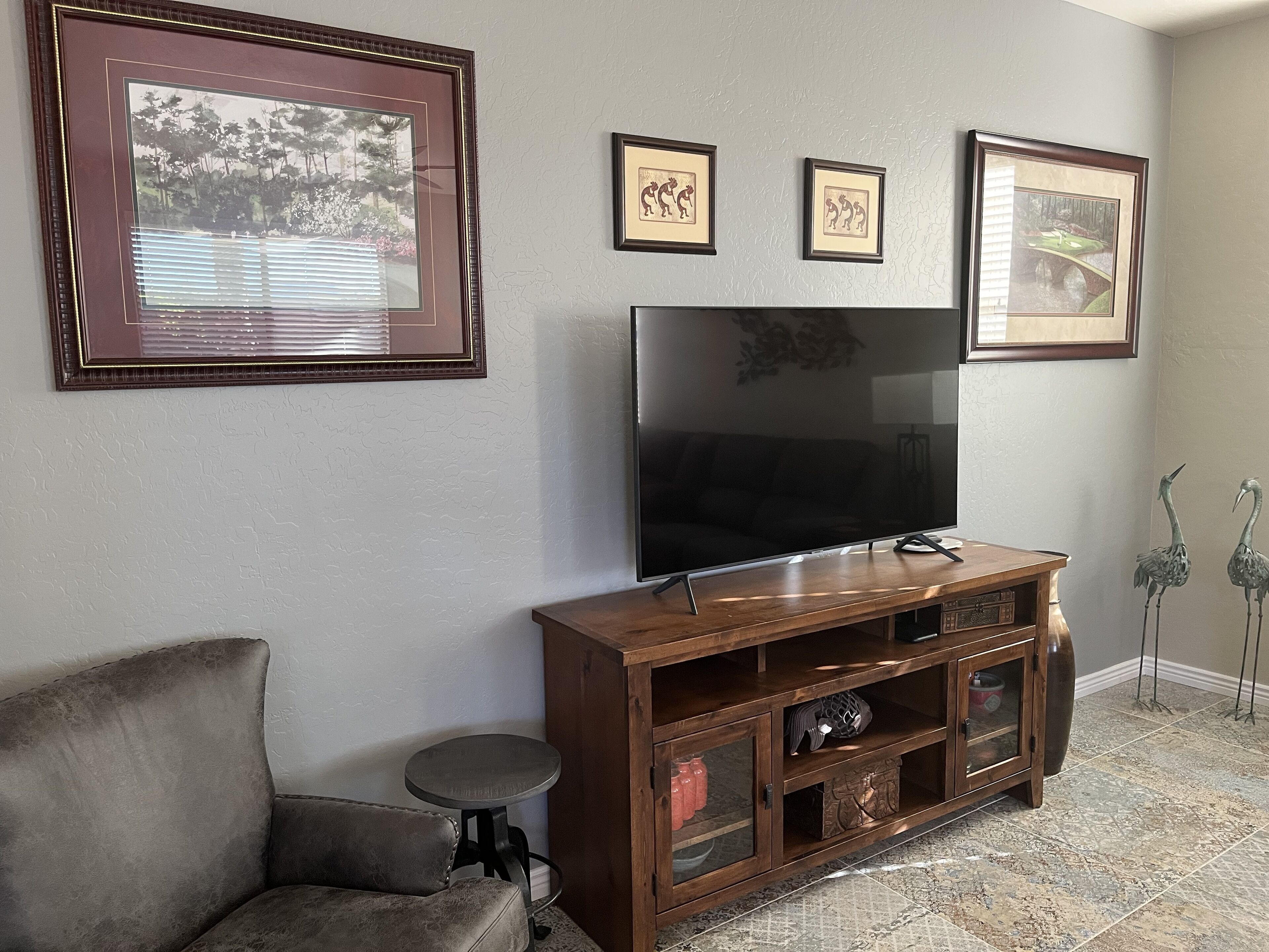 Una sala de estar con un televisor de pantalla plana grande sobre un soporte de madera, un sofá gris y tres cuadros en la pared.
