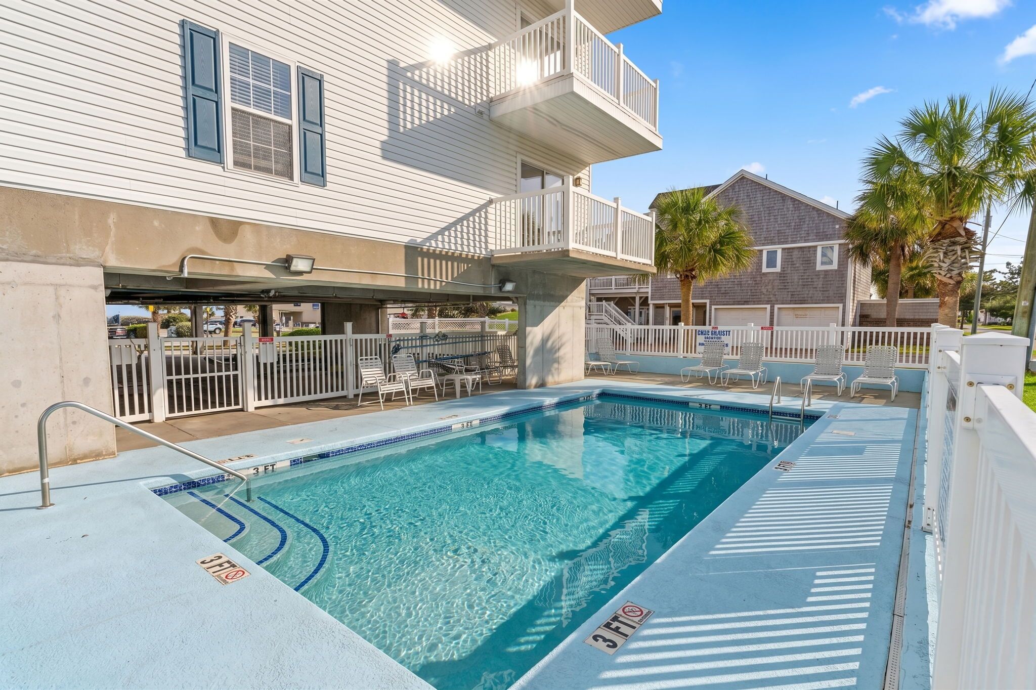 A swimming pool with a 3ft depth marker, a covered area with chairs, and a building with a balcony.