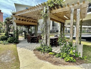 A wooden pergola structure with a table and chairs underneath, surrounded by greenery and a paved area. - Beautiful 2nd Floor Condo located on the Beach with Views of The Pass (Orange Beach)