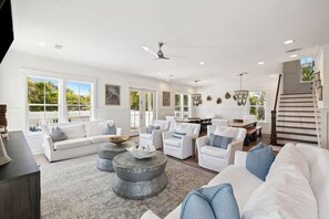 A spacious living room with a white sofa, armchairs, and a coffee table. There is a staircase leading to another level, and large windows providing natural light.