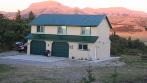 A two-story house with a green roof and a garage, surrounded by a grassy area and mountains in the background. - Private Retreat in the Rocky Mountain Front with a Million Dollar View  (Choteau)