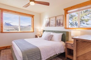 Un dormitorio con una cama grande, dos mesitas de noche, un escritorio y un ventilador de techo. Hay tres ventanas que ofrecen vistas al exterior.