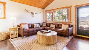 Un salon chaleureux avec un canapé d’angle en cuir marron, une table basse en bois et une grande fenêtre donnant sur des arbres et des montagnes.