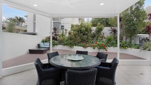 Une terrasse couverte avec une table ronde en verre, entourée de chaises en osier, d’un petit canapé et de diverses plantes et fleurs en pot.