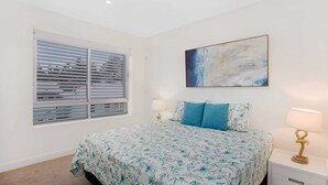 A bedroom with a bed, a window with blinds, a nightstand with a lamp, and a painting on the wall. - Rainbow Bay Villa - Spacious and Luxurious (Coolangatta)
