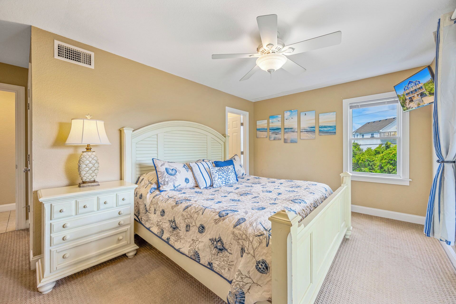 A bedroom with a bed, nightstand, lamp, ceiling fan, window, and artwork on the wall.