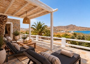 A covered outdoor seating area with stone flooring, lounge chairs, and a view of the sea and hills.