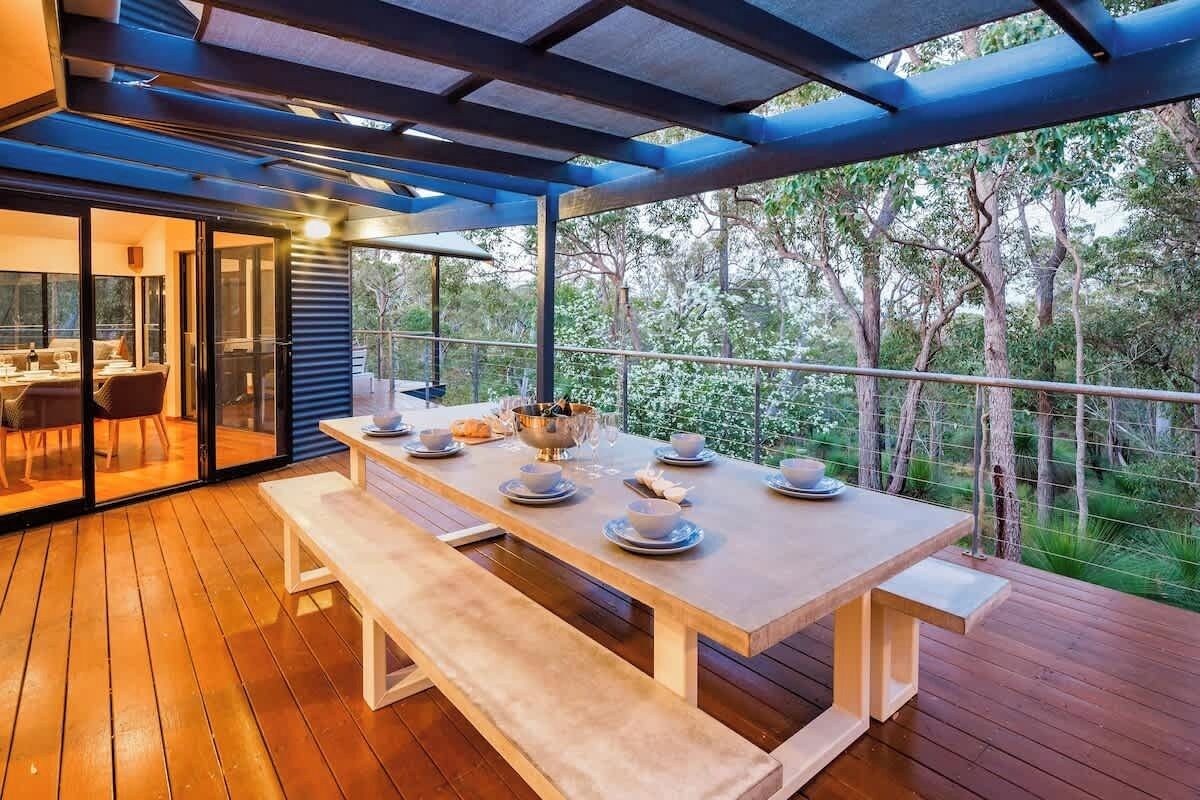 An outdoor dining area with a wooden table and benches, surrounded by a glass railing with a view of trees.