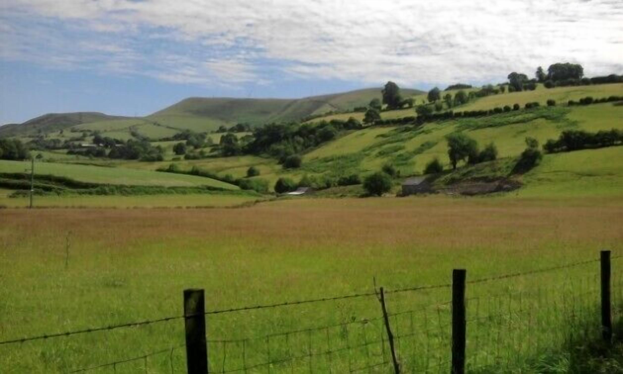 Ländliche Landschaft mit sanften Hügeln, einem eingezäunten Feld und einem kleinen Gebäude in der Ferne.