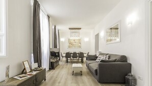Ein modernes Wohnzimmer mit einem grauen Sofa, einem Esstisch mit Stühlen, einem kleinen Hocker und einer Kommode mit Schubladen.