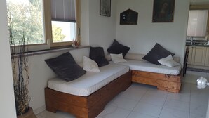 Ein gemütliches Zimmer mit einer hölzernen Sitzbank und einer gepolsterten Bank mit Kissen, einem Fenster mit Jalousien sowie einer Küchenzeile im Hintergrund.