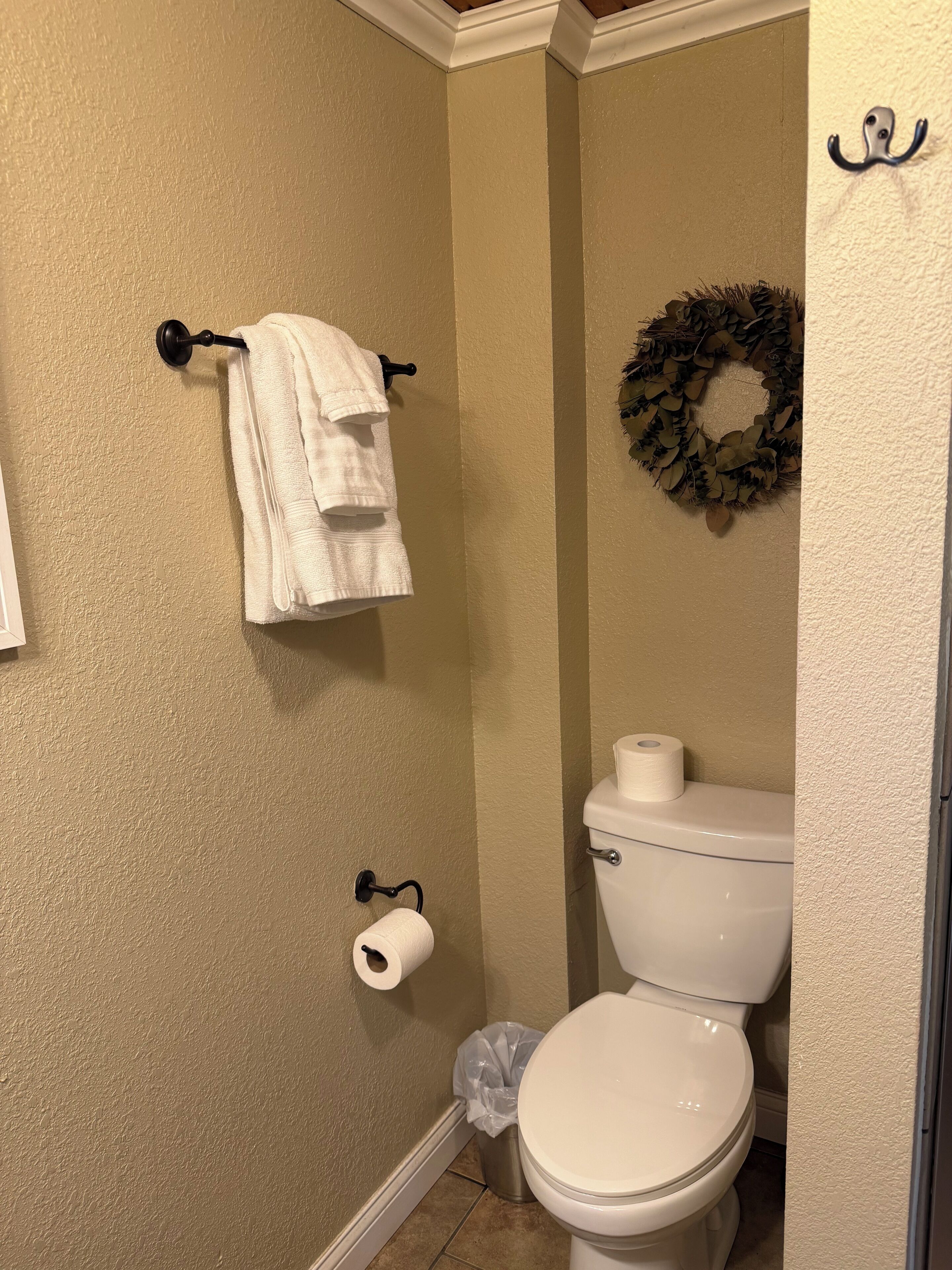A bathroom with a toilet, a towel rack with two towels, a toilet paper holder, a trash can, and a wreath on the wall.