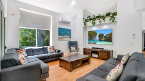 A modern living room with a grey sofa set, a wooden coffee table, and a TV on a wooden stand. There are two windows with a view of greenery outside. - 7 Naiad Court - Rainbow Shores. Swimming Pool and Walk To Beach. Aircon. Wi-Fi. (RAINBOW BEACH)
