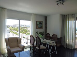 A living room with a sofa, dining table with chairs, a painting, and a view of palm trees and buildings through large windows.