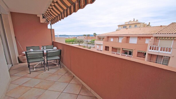 A balcony with a table and chairs, a striped awning, and a view of buildings and a body of water. - Apartment for 4 people – Sea view – Air conditioning – Downtown – Sainte Maxime (STE MAXIME)