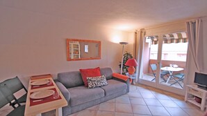 A living room with a grey sofa, a small table with plates and glasses, a red lamp, and a sliding glass door leading to an outdoor area with a table and chairs. - Apartment for 4 people – Sea view – Air conditioning – Downtown – Sainte Maxime (STE MAXIME)