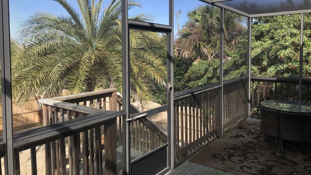 A covered outdoor area with a wooden deck, glass walls, and a view of palm trees and greenery.