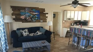 A living room with a blue sofa, a coffee table, and a painting of a turtle on the wall. There is a kitchen area with a bar and stools.