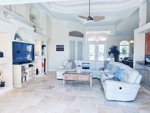 A spacious living room with a white leather sectional sofa, a wooden coffee table, a flat-screen TV, and a ceiling fan.