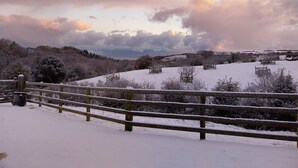 Schneebedeckte Landschaft mit Holzzaun, Bäumen und einer entfernten Hügellandschaft.