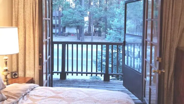 Un dormitorio con cama, mesita de noche, lámpara y vistas a una terraza y un bosque a través de una puerta abierta.