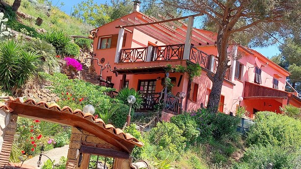 Una casa de dos pisos con fachada roja, un balcón y un jardín con diversas plantas y flores.