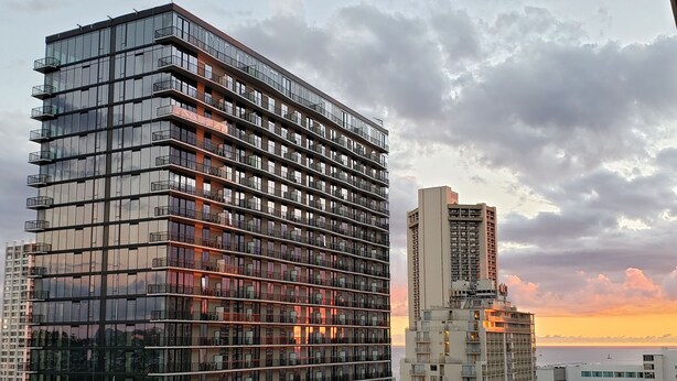 A high-rise building with multiple floors and balconies, surrounded by other buildings and palm trees.