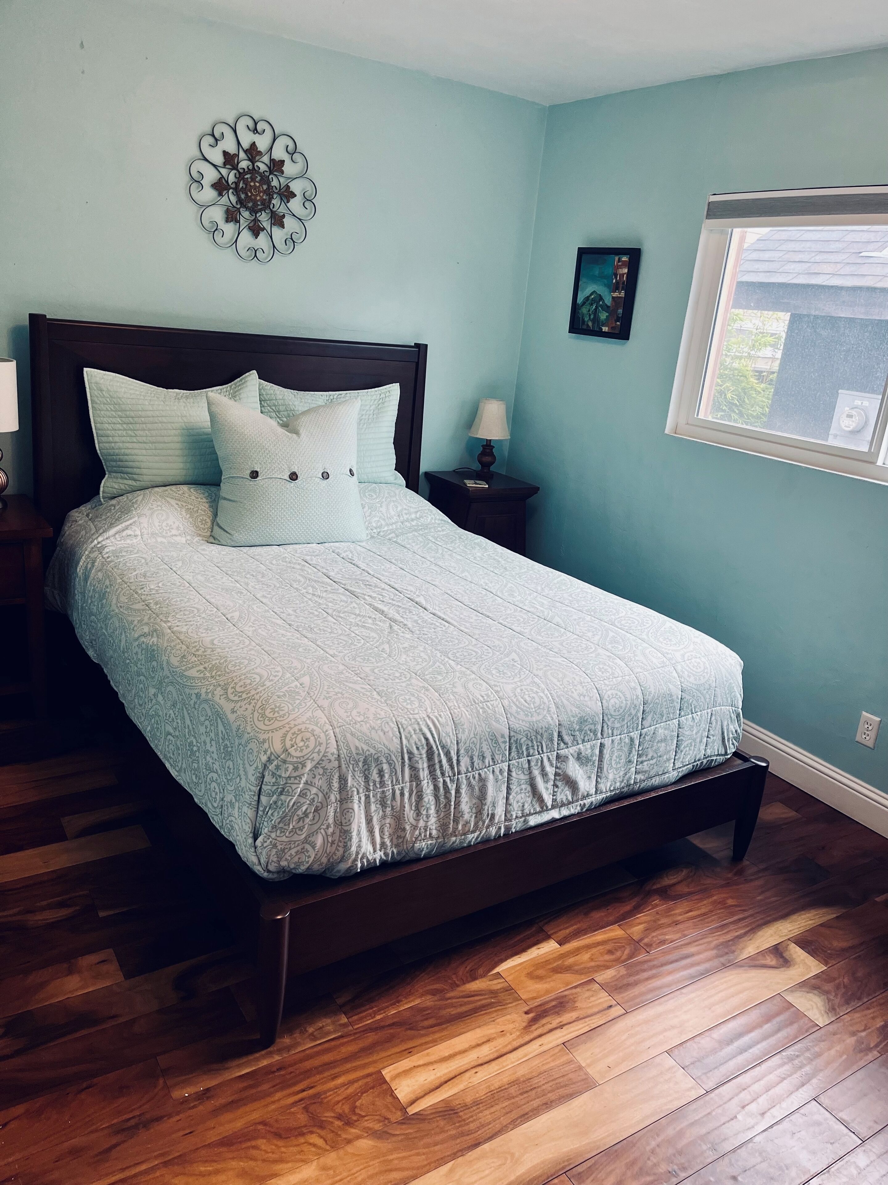 A bedroom with a bed, wooden flooring, a nightstand, a lamp, a wall decoration, and a window.